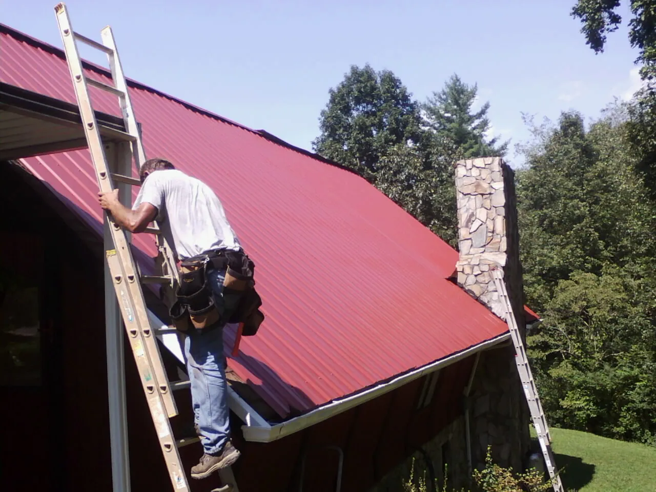 Roofer on a ladder beside a red metal roof and stone chimney — Soffit & Fascia Repair by Roofing Brothers in Jenners